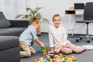 cute girl sitting on floor and looking at camera while boy  playing with multicolored wooden blocks in apartment
