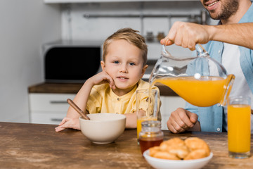 cropped view of smiling father pouring orange juice in glass and cute boy looking at camera