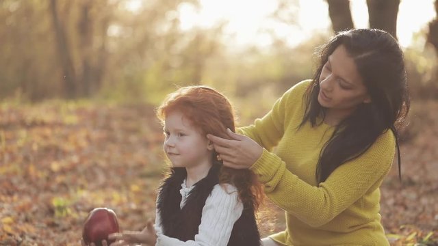Beautiful young mother and her cute redhead daughter have fun in the autumn park. The mother takes care of her daughter, braids her hair