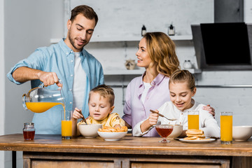 smiling woman looking at husband pouring orange juice in glass while children eating oatmeal