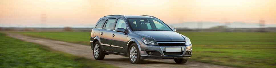 Gray silver new shiny car moving on empty rural road on blurred green meadows and clear foggy sky...