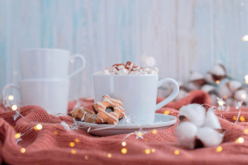 Coffee cup with marshmallow and stars cookies on warm knitted coral blanket, christmas lights and cotton flowers.