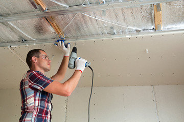 Young man in goggles fixing drywall suspended ceiling to metal frame using electrical screwdriver...
