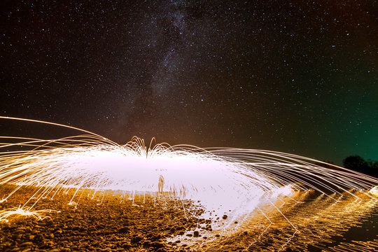 Light painting art concept. Spinning steel wool in abstract circle, firework showers of bright yellow glowing sparkles in fountain form on river bank on blue night starry sky copy space background.