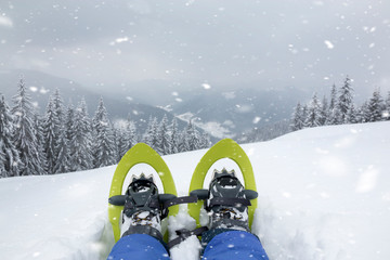 POV view of legs and trekking shoes with snowshoes on background of mountains and snow forest. The traveler sits on top of the mountain and looks at the horizon