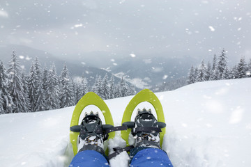 POV view of legs and trekking shoes with snowshoes on background of mountains and snow forest. The...