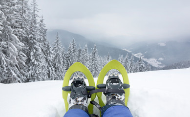 POV view of legs and trekking shoes with green snowshoes on background of mountains and snow forest.