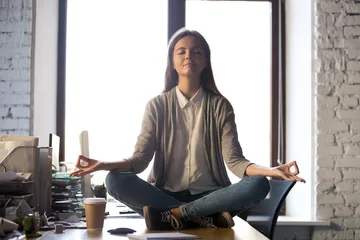 Gordijnen Serene calm business woman sit on office desk taking break for meditation, mindful employee doing yoga exercise in lotus pose for relaxation at workplace, no stress relief, balance at work concept © fizkes