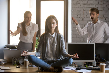 Calm serene employee meditating in office ignoring not listening to annoying colleagues, funny...