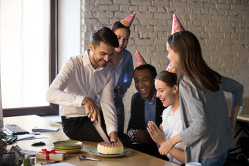 Friendly multi-ethnic employees in party hats cutting cake celebrating excited colleague happy birthday together, office workers congratulating coworker making corporate surprise having fun concept