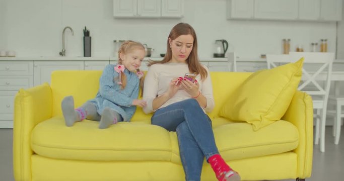 Attractive single mother busy with cellphone phubbing her adorable joyful elementary age daughter in domestic room. Lovley little girl needs attention of addicted mum using smart phone device at home.