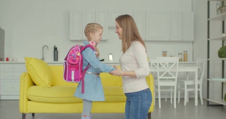 Caring mother preparing and cheering up cute little daughter with backpack before first day of school in domestic room. Affectionate mum kissing goodbye schoolgirl as she leaves for primary school.