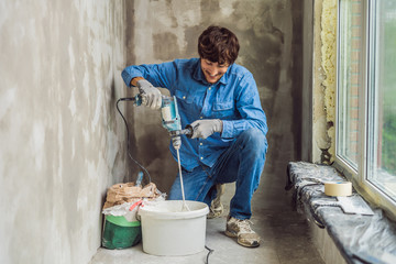 Young male painter kneads putty with water in a bucket using a hand-held mixer for building mixes