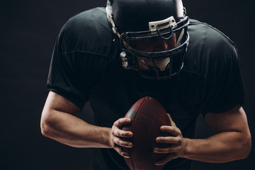 American football player with black helmet and armour running in motion, holding ball, getting ready to score a goal, close up shot over dark background - Powered by Adobe