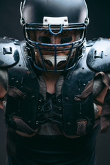 Portrait of sexy american football player wearing protective armour on shirtless torso, looking confidently, ready to tear apart everybody who bothers to win, isolated over dark studio background.