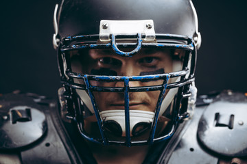 Headshot of handsome american football team quarterback wearing protective armour on shirtless torso, looking confidently through his helmet, isolated over dark studio background.