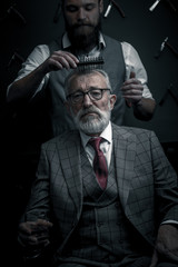 Barber making male haircut. Hairdresser cutting hair of his rich client, dressed in expensive stylish man s suit of clothes and holding a glass of alcoholic beverage
