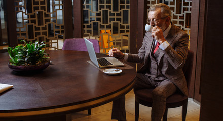 Portrait of mature wealthy man in years having coffee break at his private cabinet, home parlor, reading business article in newspaper and following to the latest news in laptop screen