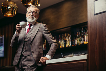 Portrait of mature old-aged successful writer ot creative designer man drinking a cup of tea after dinner in cozy twilight interior of fashionable cafe