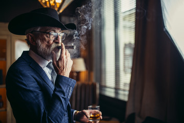 Elegant senior gentleman wearing spectacles and grey-haired beard standing at pub in wide-brimmed hat with glass of alcohol drink and cigar. Elderly man dressed in tailored classy suit and tie. - Powered by Adobe