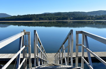 Wooden pier with stairs on a river with beach and forest. Water reflections, sunny day, blue sky. Galicia, Spain.