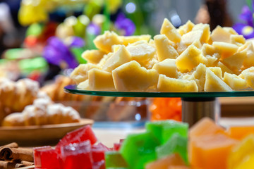 Closeup of stacks of small cubes of green, red, yellow, orange marmalade, glass round plate with hard parmesan cheese and cinnamon sticks in crystal glass on rectangular wooden plate on catering table