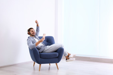 Handsome young man listening music in armchair indoors. Space for text