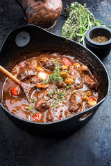 Traditionell geschmorte deutsche Schweinebäckchen in brauner Rotwein Sauce  mit Pilzen und Karotten als closeup in einem gußeisernen Topf
