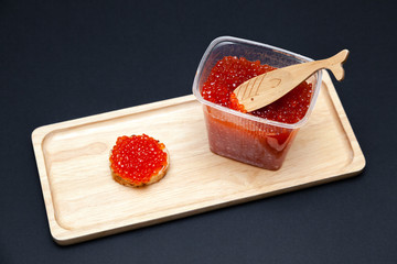 An open plastic jar of red salmon caviar with a fish-shaped spoon and a gourmet snack round sandwich on a rectangular wooden plate on a black background. Concept useful delicacy, seafood diet