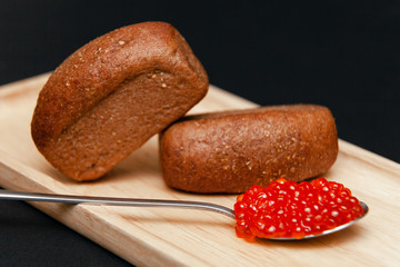 Closeup macro two small pieces black bread with cumin and metal spoon with slide of red salmon caviar gourmet snack on rectangular wooden plate on black background. Concept healthy seafood appetizer