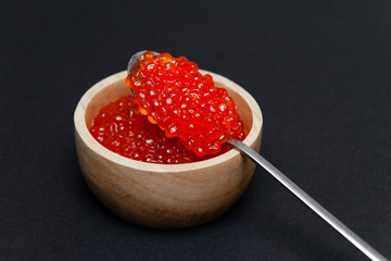 Closeup macro saucer or bowl with slide of fresh tasty red salmon caviar and metal spoon inside on black background. Concept gourmet snack seafood appetizer