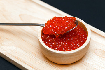 Closeup macro saucer with slide of red salmon caviar and metal spoon on rectangular wooden plate on black background. Concept gourmet snack seafood appetizer