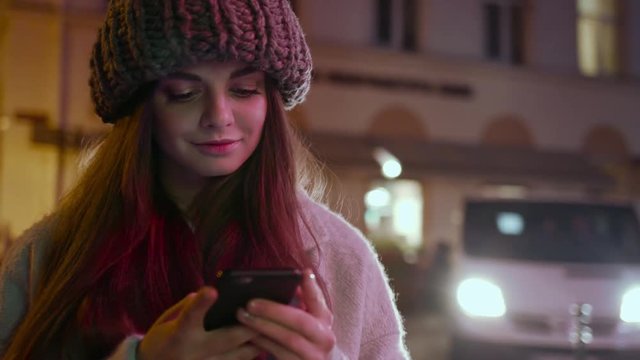 Young smiling woman with hat use of smart phone stand in the winter time at night light portrait street smart travel city evening girl message mobile online urban close up slow motion