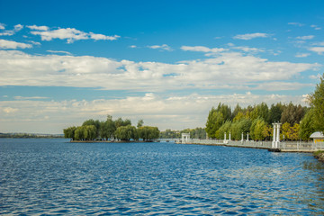 Ukrainian city park outdoor lake waterfront shoreline space for rest and walking in weekend time with view on small island and bridge on background and vivid blue water surface on foreground 