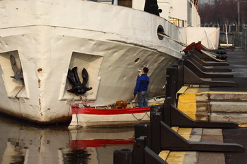 Maintenance, repair of vessel - sailor on the safety boat paints the board of the river ship at the pier