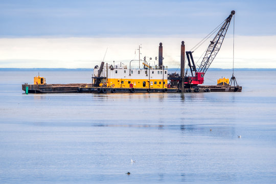 An old dredging barge in the ocean. Barge is yellow and white with a red crane on the deck. Scoop is being lowered into the water. Dark overcast sky.