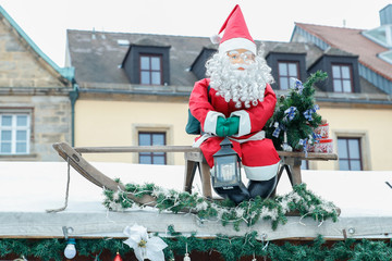 Weihnachtsmann mit grünen Handschuhen und Licht in der hand auf einem Schlitten der mit Tannenbaumästen geschmückt ist auf dem Christkindlmarkt in Bayreuth.