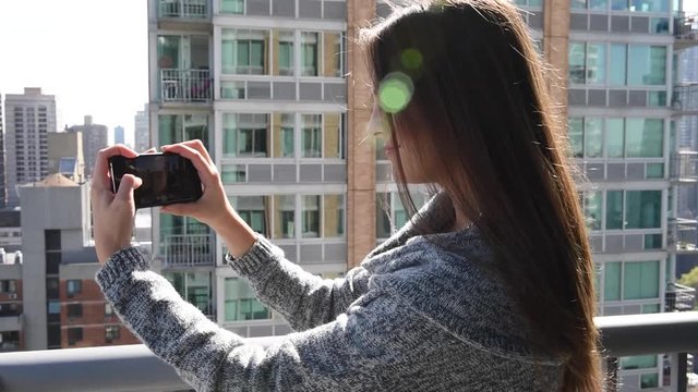 Young millennial beautiful woman or tourist taking photograph with mobile cellphone smartphone device of skyline in New York view from her apartment. Girl to share pic on social media photo site.
