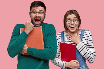 Fototapeta na wymiar Studio shot of overjoyed woman and man clench fists with happiness, feels glad to graduate from university, holds textbooks, gaze at camera, open mouth widely, isolated over pink background.
