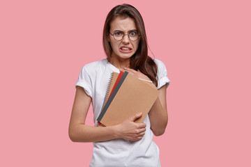 Displeased college student lady has unhappy facial expression, clenches teeth, carries spiral notebooks, has no inspiration for work, isolated over pink studio wall. Youth and studying concept