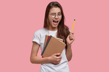 Indoor shot of happy young Caucasian female genius gets good idea, blinks eye, holds pencil, carries notebooks, has fun indoor, dressed in white casual t shirt, isolated over pink background