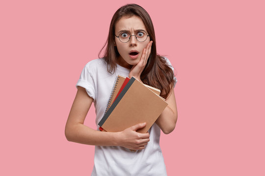 Waist up shot of stupefied Caucasian woman with frightened expression, opens mouth widely, holds notepads, dressed in white casual t shirt, stares through round spectacles, models over pink wall
