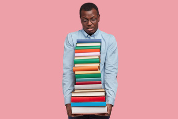 Indoor shot of displeased African American scientist or student carries heavy pile of textbooks, wears elegant shirt, models over pink studio wall, realizes how much he should learn. Education