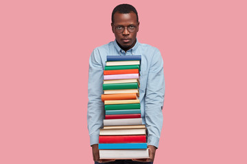 Indoor shot of serious dark skinned guy looks with dissatisfied expression, has many books to read, dressed in elegant clothes, prepares for writing course paper, isolated over pink background