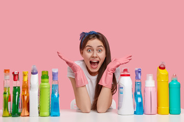 Horizontal shot of amazed overjoyed woman spreads hands, wears protective gloves, sits at table with many detergents, advertises good cleaning supplies for tidying up, isolated over pink background