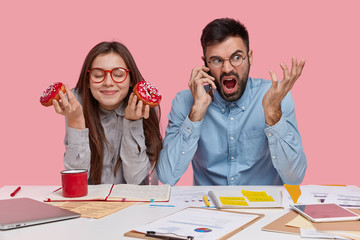 Photo of pleased lady wears red rim spectacles, eats delicious donuts, sits near her male partner who speaks over smart phone with angry expression, surrounded with notepads, isolated on pink