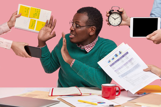Oh no, not now! Stressed dark skinned man has much work in office, asked by many people at one time who remind about preparing business project, hold alarm clock, notepad with records, sits at desktop
