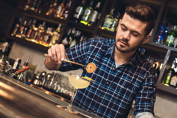 Young bartender standing at bar counter decorating cocktail with orange concentrated
