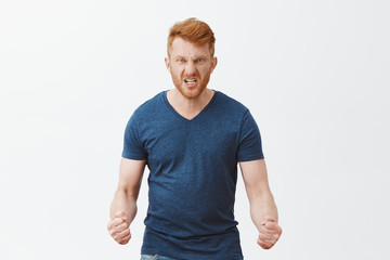 Fototapeta premium Guy will show how anger looks. Portrait of pissed and angry dangerous redhead guy with outraged expression, wrinkling nose, frowning and clenching fists dangerously, feeling hate over gray wall
