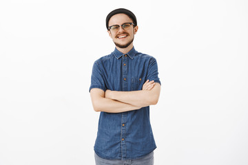 Ambitious creative and handsome friendly-looking mature guy with moustache in black beanie and glasses holding hands crossed on chest with confident and self-assured smile posing over gray background